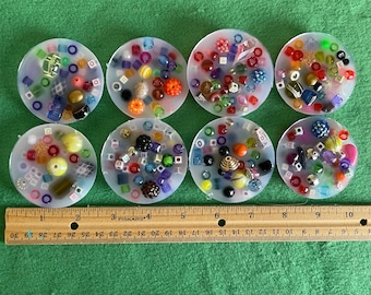 May include: Eight clear round coasters with colorful beads and small objects inside. The coasters are arranged in two rows of four. A ruler is visible at the bottom of the image.