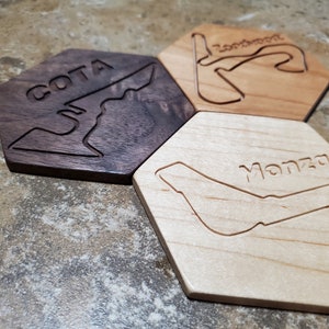 May include: Three hexagonal wooden coasters with engraved race track designs. One is dark brown with "COTA", another is light brown with "Zandvoort", and the third is light wood with "Monza".