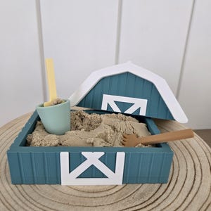 May include: A teal barn-shaped sandbox filled with beige kinetic sand. The set includes a light green bucket with a yellow shovel, a brown rake, and a white barn roof piece. The sandbox features white cross-brace detailing on the front panel.