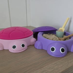 Puede incluir: Dos recipientes con forma de tortuga, uno rosa claro con tapa de caparazón rosa oscuro y otro morado con tapa a juego. El recipiente morado está abierto, lleno de arena, y contiene una pequeña pala amarilla clara y una herramienta de madera.