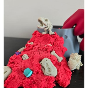 May include: A pile of bright red kinetic sand featuring several light gray plastic dinosaur skeleton figures, including a T-Rex, and small faceted purple and blue plastic gems. A gray plastic mold container sits behind the sand pile.