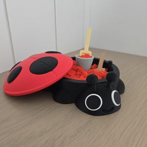 May include: A black ladybug-shaped container with a red lid featuring black spots. The container is filled with orange kinetic sand and includes a small light-colored plastic bucket and a matching shovel tool resting inside the sand.
