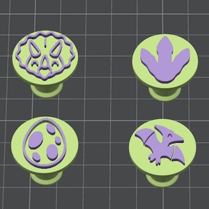 May include: Four circular light green stamps with purple raised designs. The designs include a triceratops head, a three-toed dinosaur footprint, a spotted dinosaur egg, and a flying pterodactyl. Each stamp has a cylindrical handle base.