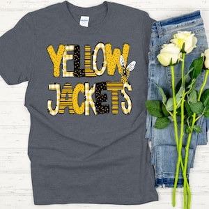 May include: A dark heather gray short-sleeve t-shirt featuring the text YELLOW JACKETS in a bold, blocky font. The letters are filled with yellow and black patterns including stripes, polka dots, and gingham, with a small bee illustration.
