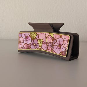 May include: A dark brown hair claw clip with a rectangular floral design. The design features pink and purple flowers with green leaves, outlined in gold. The clip is made of a matte material.