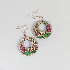 May include: A pair of wooden hoop earrings with floral designs. Each earring features a circular wooden base with a central hole, adorned with pink, white, and green painted flowers. The earrings have rose gold-colored ear wires.