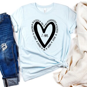 May include: A light blue t-shirt with a black heart graphic and the text "I am" inside the heart. The heart is surrounded by a circle of words including "amazing", "beautiful", "kind", "wise", "courageous", "talented", "brave", "unique", "enough", "smart", and "me".