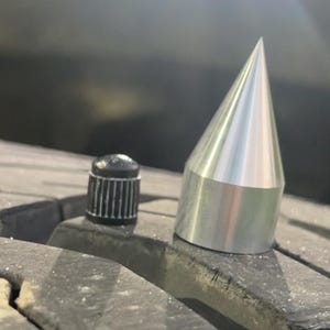 May include: A silver metal tire spike and a black tire valve cap are shown on a tire. The spike has a conical shape with a flat base. The valve cap is cylindrical with vertical grooves.