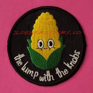 May include: A black and white embroidered patch featuring a cartoon corn cob with a face and the text "The lump with the knobs".