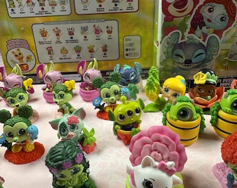 May include: A collection of colorful Disney character figurines, including Mickey Mouse, Stitch, and Marie, displayed on a pink surface. The figurines are part of a "In Full Bloom" series, with a box featuring character illustrations in the background.
