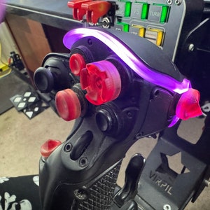 以下が含まれることがあります： 赤色のボタンと紫色のライトが付いた黒いジョイスティック。ジョイスティックには複数のボタンとスイッチがあり、金属製のベースに取り付けられています。画像はジョイスティックのクローズアップで、そのデザインと機能を強調しています。