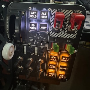 Peut inclure: Un panneau noir avec divers interrupteurs et boutons, y compris un levier de train d'atterrissage, des interrupteurs de trim et un interrupteur de verrière. Le panneau est étiqueté avec du texte tel que "ATT HOLD", "HDG HOLD" et "FCS RESET".