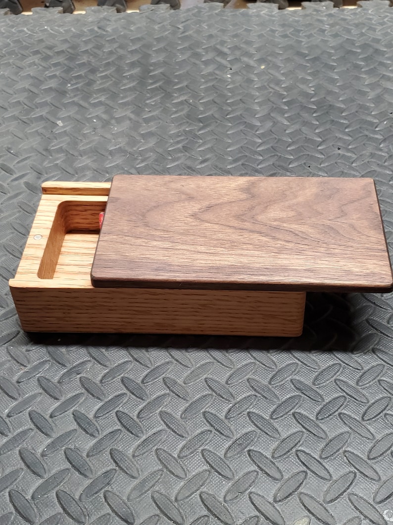 May include: A wooden box with a hinged lid. The box is made of light brown wood and has a dark brown wood lid. The lid is open and the inside of the box is visible. The box is sitting on a gray textured surface.