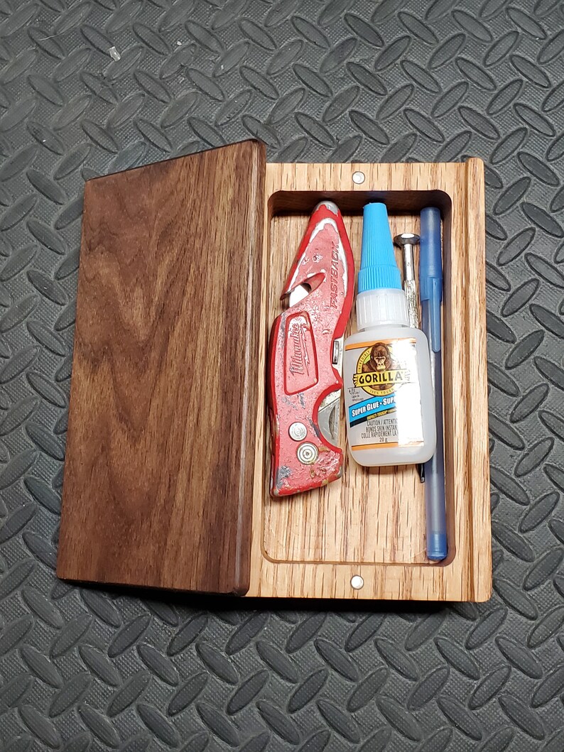 May include: A wooden box with a hinged lid, containing a red utility knife, a blue bottle of Gorilla Super Glue, a silver metal screw, and a blue pen.