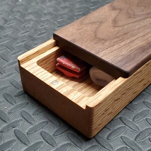 May include: A wooden box with a hinged lid, made of walnut and oak wood. The box is open, revealing a red object and a small, white object inside.