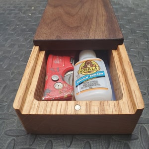 May include: A wooden box with a sliding lid, containing a red utility knife and a bottle of Gorilla Super Glue. The glue bottle is white with a black label and the text "Gorilla Super Glue - Super Colle" in black and blue.