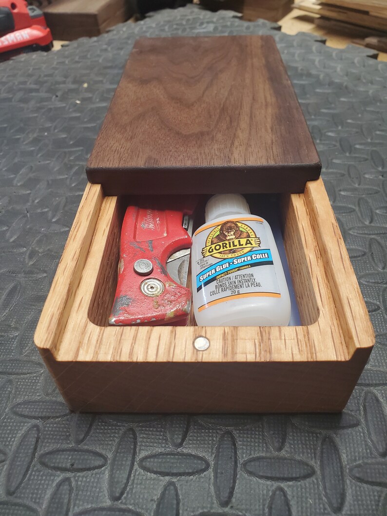 May include: A wooden box with a sliding lid, containing a red utility knife and a bottle of Gorilla Super Glue. The glue bottle is white with a black label and the text "Gorilla Super Glue - Super Colle" in black and blue.