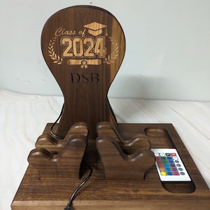 May include: A wooden phone charging station with a graduation cap design. The station has three phone holders and a remote control for the LED lights. The station is engraved with "Class of 2024" and "DSB".
