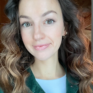 May include: A woman wearing a dark green corduroy jacket over a white shirt. The jacket has silver buttons and a pocket on the chest. She has brown hair with blonde highlights and is wearing small gold hoop earrings.