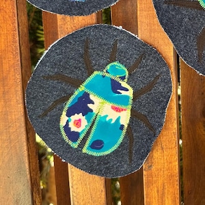 Op de afbeelding: Vier decoratieve onderzetters met een keverontwerp. Elke onderzetter heeft een donkergrijze achtergrond met een kleurrijke keverillustratie in tinten blauw, turquoise, roze en wit. De onderzetters zijn geplaatst op een houten oppervlak.