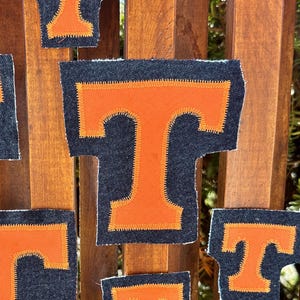 May include: A collection of orange and navy blue fabric patches in the shape of the letter "T". The patches are arranged on a wooden background. The patches vary in size and are likely for crafting or apparel.