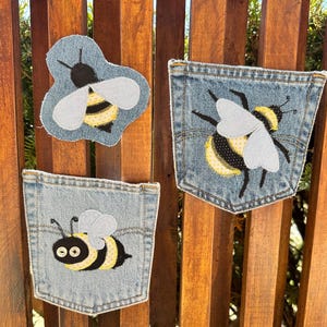 May include: Three denim patches featuring bee designs. One is a bee shape, and the other two are jean pockets. The bees are black, yellow, and white, with embroidered details. The patches are displayed against a wooden background.