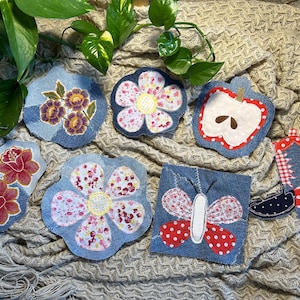 May include: A collection of denim coasters in various shapes, including flowers, an apple, a butterfly, and a cowboy boot. The coasters feature colorful fabric appliqués and decorative stitching. The background is a textured, neutral-toned fabric.