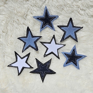 May include: A collection of seven star-shaped fabric patches arranged in a cluster. The patches feature various shades of blue denim, dark navy fabric, and solid white material, each outlined with visible black stitching along the edges.