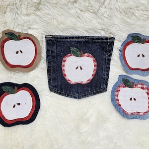 May include: A dark blue denim pocket featuring a central apple applique, surrounded by four individual apple-shaped fabric coasters or patches. Each apple design consists of a white center, a red border, and a small green leaf at the top.