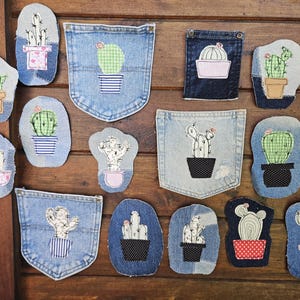 May include: A collection of denim fabric patches featuring embroidered cactus designs in various pots. The patches include individual shapes and detached denim pockets, showcasing green, white, and blue patterns with stitched details on blue fabric.