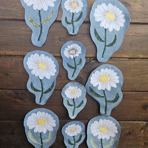 May include: A collection of ten fabric patches, each featuring an embroidered white daisy with a yellow center on a light blue denim-like background. The patches vary in size and shape, with green embroidered stems and leaves on each flower.