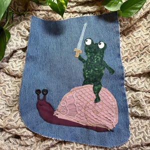 May include: A denim patch featuring an applique design of a green frog holding a sword, riding a pink snail. The snail has a lace shell and a maroon body. The frog and snail are set against a blue denim background.