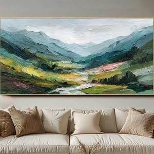 May include: An abstract landscape painting in a gold frame. The artwork shows a valley between mountains, in shades of green, blue, yellow, and pink. The painting hangs above a beige sofa with brown cushions.