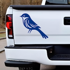 May include: A blue bird decal applied to the white tailgate of a pickup truck. The graphic features a stylized bird in profile, facing left, with clean lines and a solid blue color. The decal is positioned centrally on the flat white surface.