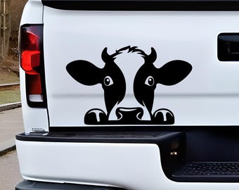 Könnte beinhalten: Ein schwarz-weißer Vinyl-Aufkleber eines Kuhgesichts, das über eine Kante schaut, angebracht auf der weißen Heckklappe eines Pick-ups. Die Grafik zeigt eine stilisierte Kuh mit großen Augen, Ohren und Hufen am unteren Rand des Designs.