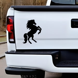 May include: A black silhouette of a rearing horse is affixed to the white tailgate of a truck. The horse is depicted with a flowing mane and tail, and is in a dynamic pose. The truck's taillight is visible on the left side.