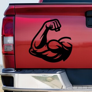 May include: A black vinyl decal featuring a stylized graphic of a flexed bicep arm, applied to the red tailgate of a pickup truck. The design shows a solid black silhouette of a muscular arm with a clenched fist, positioned on the flat surface.