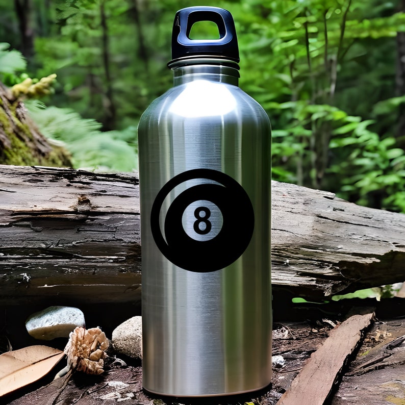 K&ouml;nnte beinhalten: Eine Edelstahl-Wasserflasche mit schwarzem Deckel und Griff. Die Flasche hat eine schwarz-wei&szlig;e Grafik einer Acht-Ball-Kugel. Der Hintergrund zeigt eine nat&uuml;rliche Au&szlig;enszene mit B&auml;umen und Holz.