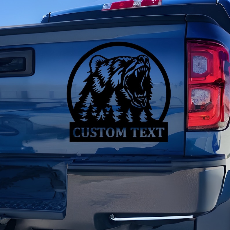 K&ouml;nnte beinhalten: Ein schwarzer, kreisf&ouml;rmiger Aufkleber auf der blauen Heckklappe eines Pick-ups. Das Design zeigt die Silhouette eines br&uuml;llenden B&auml;ren mit einem integrierten Kiefernwald im unteren Bereich und dem Schriftzug CUSTOM TEXT in fetter Schrift.