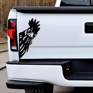 May include: Black rooster decal on the white tailgate of a truck. The rooster is a stylized silhouette, with a detailed comb and beard. The decal is positioned on the right side of the tailgate, near the taillight.