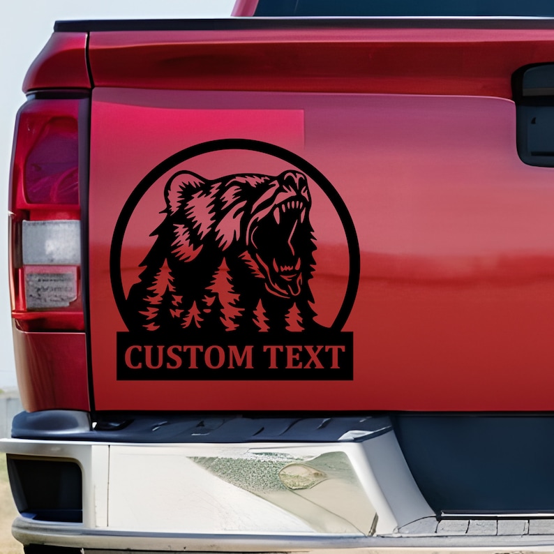 K&ouml;nnte beinhalten: Ein schwarzer, kreisf&ouml;rmiger Aufkleber auf der Heckklappe eines roten Pickups. Das Design zeigt die Silhouette eines br&uuml;llenden B&auml;ren mit einem integrierten Tannenwald im Fell, platziert &uuml;ber einem rechteckigen Banner mit dem Text CUSTOM TEXT.