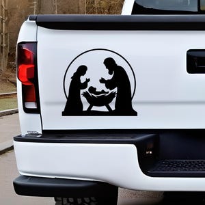 May include: Black silhouette decal depicting the Nativity scene, featuring Mary, Joseph, and baby Jesus in a manger, enclosed within a circular frame. The decal is affixed to the tailgate of a white truck, creating a religious-themed design.