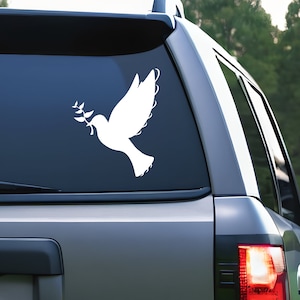 May include: White dove decal with an olive branch, applied to a vehicle window. The dove is a symbol of peace and is depicted in a simple, elegant style. The decal is a popular choice for car decoration.