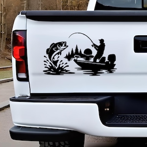 May include: A black vinyl decal applied to the white tailgate of a pickup truck. The graphic features a silhouette of a person in a boat fishing with a rod, a large fish jumping out of the water, a forest of pine trees, and a circular sun.