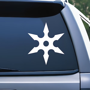 May include: White vinyl decal of a shuriken, a traditional Japanese concealed weapon. The six-pointed star design features sharp points and circular cutouts. The decal is displayed on a car window.