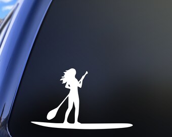 May include: A white vinyl decal sticker featuring a silhouette of a person standing on a paddleboard and holding a paddle. The design shows the figure with long hair flowing in the wind, positioned on a long, narrow board, applied to a dark surface.