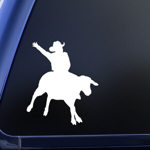 May include: White silhouette decal of a cowboy riding a bull. The cowboy is wearing a hat and has one arm raised. The bull is bucking.