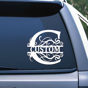 May include: A white vinyl decal applied to a vehicle rear window, featuring a large stylized letter C with intricate scrollwork patterns and a horizontal central bar containing the word CUSTOM in bold, capital, sans-serif lettering.