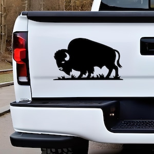 May include: A black silhouette of a bison, depicted in profile, is affixed to the white surface of a vehicle. The bison stands on a patch of grass. The image is a decal or sticker, likely for a car or truck.