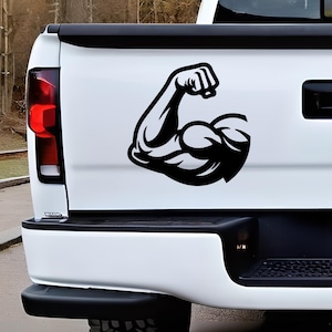 May include: A black vinyl decal featuring a stylized graphic of a flexed bicep muscle, applied to the white tailgate of a pickup truck. The design shows a solid black silhouette of a muscular arm with a clenched fist, positioned centrally.